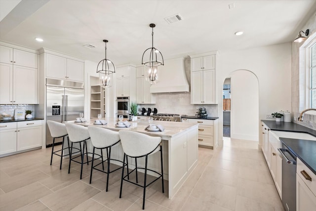 kitchen featuring appliances with stainless steel finishes, backsplash, custom range hood, a kitchen island with sink, and pendant lighting