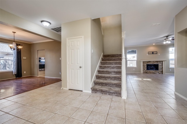 stairs featuring tile patterned floors, a stone fireplace, crown molding, and ceiling fan with notable chandelier