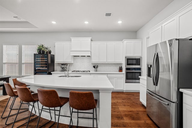 kitchen featuring a kitchen island with sink, wine cooler, decorative backsplash, white cabinets, and appliances with stainless steel finishes