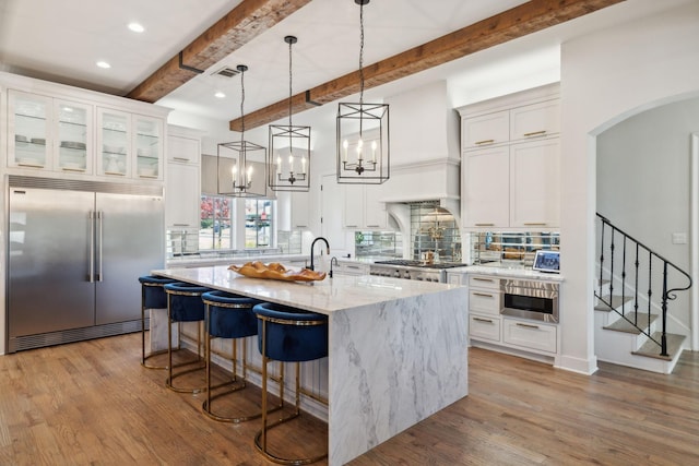 kitchen with decorative backsplash, light stone countertops, an island with sink, beam ceiling, and stainless steel appliances