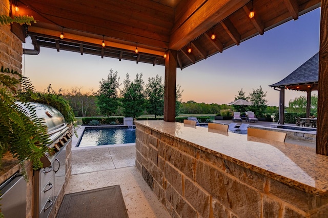 pool at dusk with a gazebo, an outdoor kitchen, and a patio