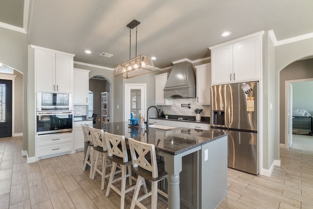 kitchen with a kitchen island with sink, premium range hood, white cabinets, decorative backsplash, and appliances with stainless steel finishes