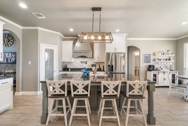 kitchen with premium range hood, hanging light fixtures, stainless steel fridge with ice dispenser, an island with sink, and white cabinetry