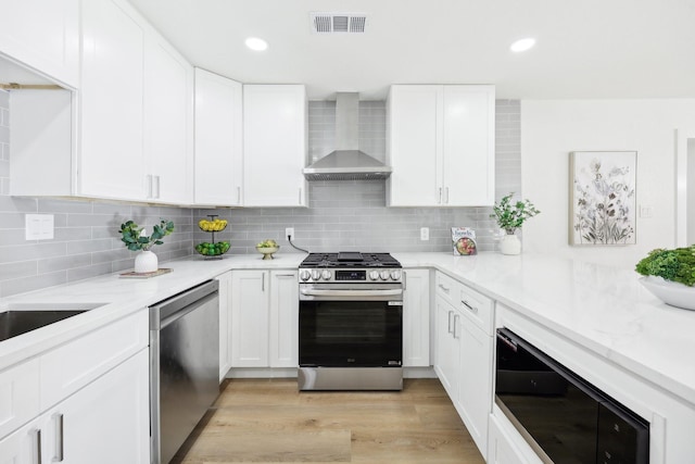 kitchen with backsplash, white cabinets, wall chimney exhaust hood, appliances with stainless steel finishes, and light hardwood / wood-style floors