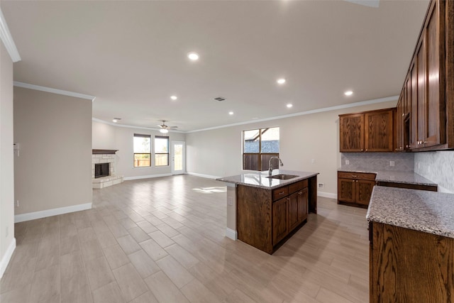 kitchen with a center island with sink, a stone fireplace, sink, ceiling fan, and light stone counters