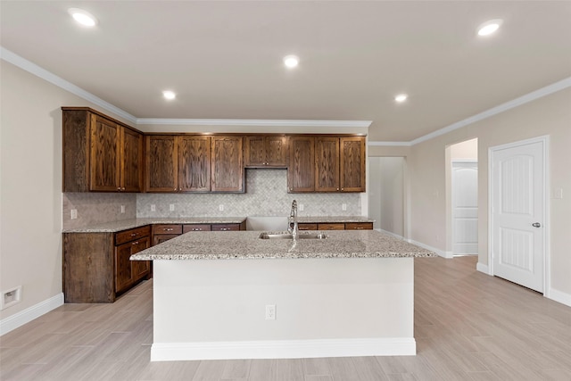 kitchen with sink, tasteful backsplash, light stone counters, an island with sink, and ornamental molding