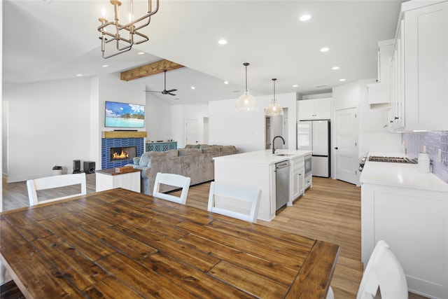 dining area with light wood-type flooring, ceiling fan with notable chandelier, sink, vaulted ceiling with beams, and a stone fireplace