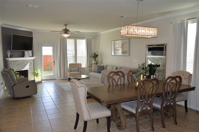 dining space with a stone fireplace, light tile patterned flooring, ceiling fan with notable chandelier, and ornamental molding