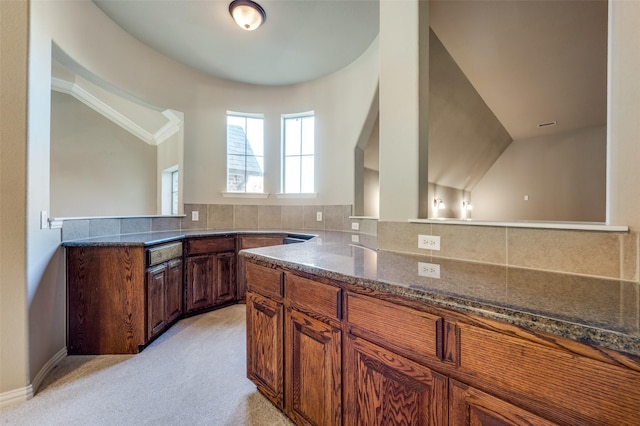 kitchen with decorative backsplash, dark stone counters, light colored carpet, vaulted ceiling, and crown molding