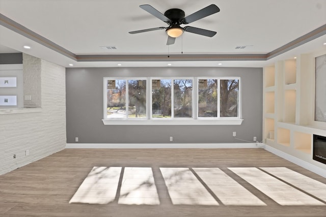 empty room with a brick fireplace, light hardwood / wood-style flooring, a raised ceiling, and ceiling fan