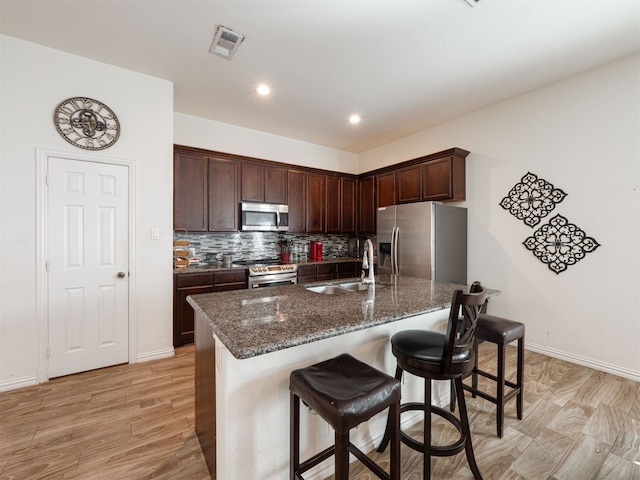 kitchen with sink, dark stone counters, a kitchen island with sink, appliances with stainless steel finishes, and light wood-type flooring