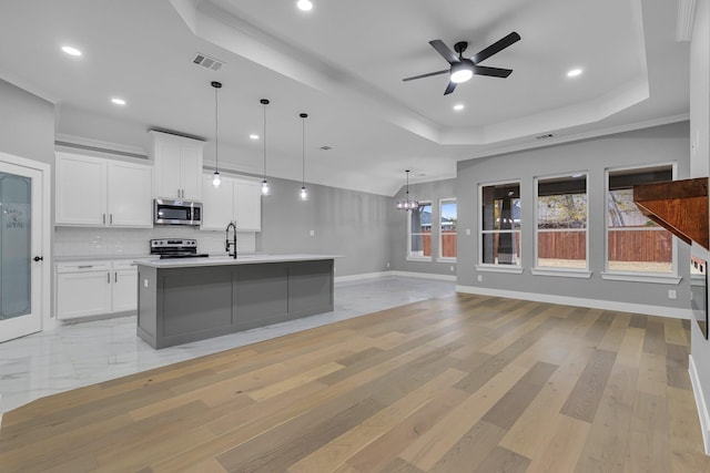 kitchen featuring appliances with stainless steel finishes, ceiling fan with notable chandelier, a raised ceiling, white cabinetry, and an island with sink