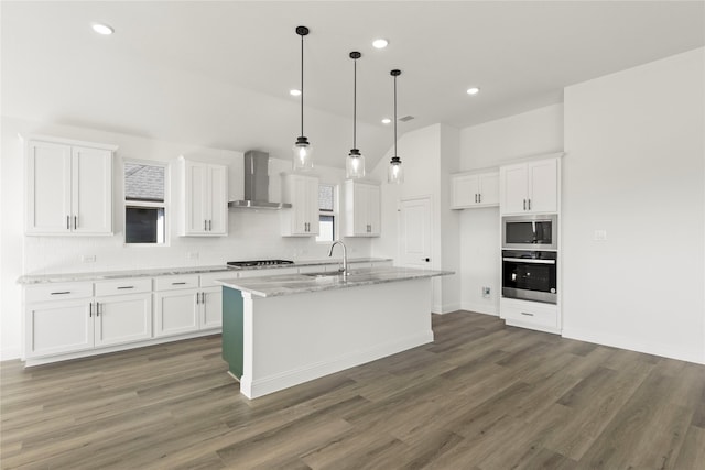 kitchen featuring wall chimney exhaust hood, built in microwave, white cabinets, oven, and an island with sink