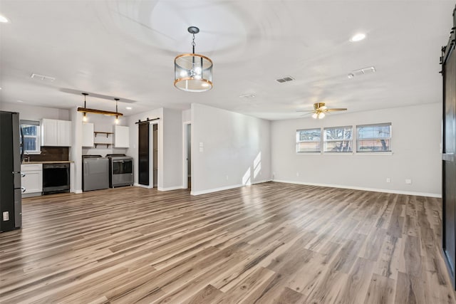 unfurnished living room with washing machine and clothes dryer, ceiling fan, a barn door, a healthy amount of sunlight, and light hardwood / wood-style flooring