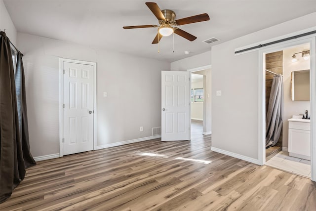 unfurnished bedroom with light hardwood / wood-style flooring, a barn door, ceiling fan, and ensuite bathroom