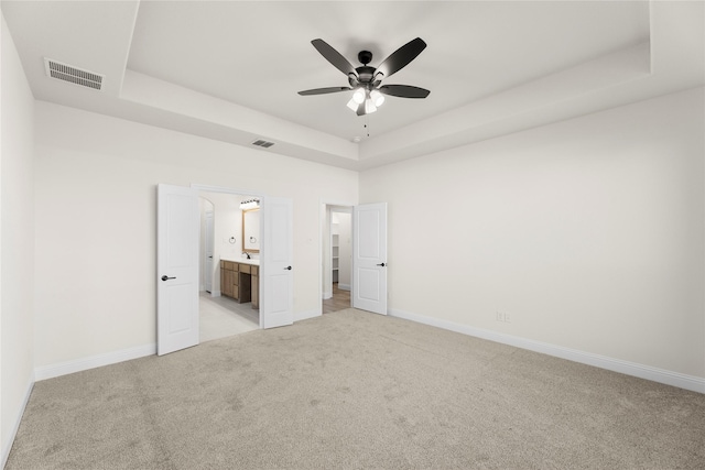 unfurnished bedroom with a raised ceiling, ceiling fan, light carpet, and ensuite bath