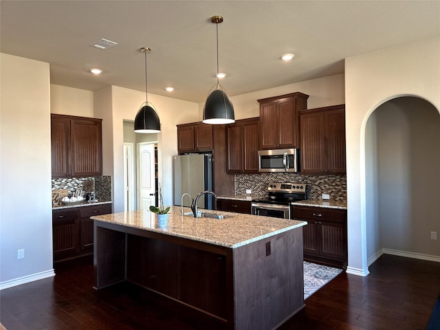 kitchen featuring appliances with stainless steel finishes, an island with sink, hanging light fixtures, light stone countertops, and dark brown cabinets