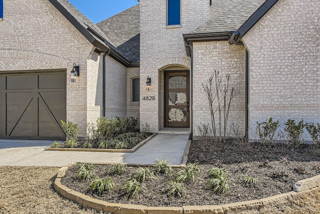 doorway to property with a garage, brick siding, and roof with shingles