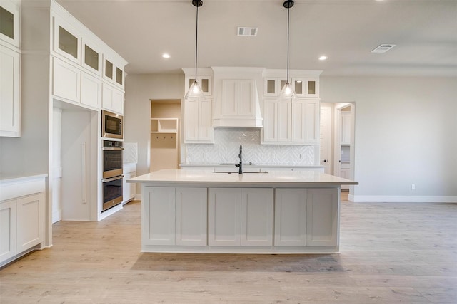 kitchen featuring premium range hood, hanging light fixtures, light hardwood / wood-style flooring, an island with sink, and built in microwave