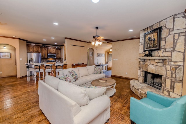 living room with dark wood-type flooring, ceiling fan, ornamental molding, and a stone fireplace