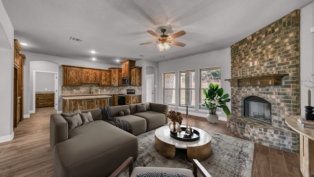 living room featuring wood-type flooring, a textured ceiling, a brick fireplace, and ceiling fan