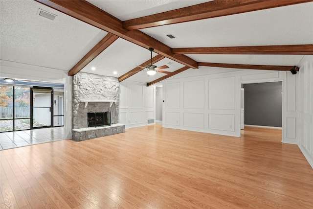 unfurnished living room featuring ceiling fan, vaulted ceiling with beams, a stone fireplace, light hardwood / wood-style flooring, and a textured ceiling