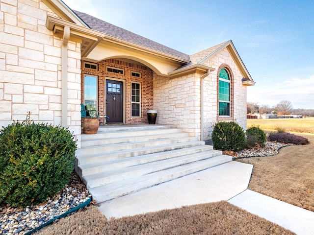 doorway to property with stone siding, brick siding, a porch, and roof with shingles