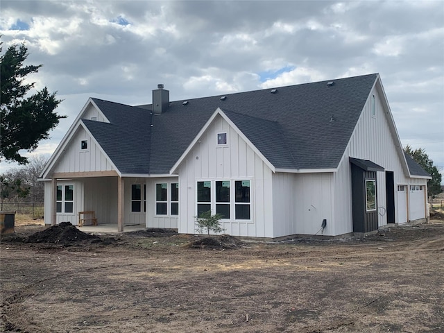 rear view of house featuring board and batten siding, roof with shingles, a patio, and a chimney