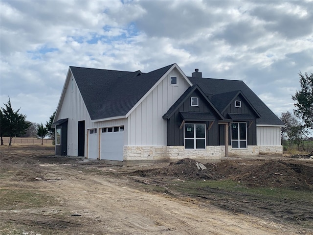 modern farmhouse with stone siding, driveway, roof with shingles, board and batten siding, and a chimney