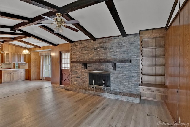 unfurnished living room with wood walls, ceiling fan with notable chandelier, vaulted ceiling with beams, a fireplace, and light hardwood / wood-style floors