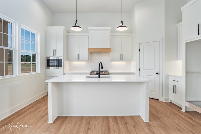 kitchen with hanging light fixtures, stainless steel microwave, light hardwood / wood-style floors, an island with sink, and white cabinets