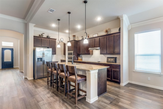 kitchen with appliances with stainless steel finishes, pendant lighting, an island with sink, dark brown cabinetry, and light stone counters