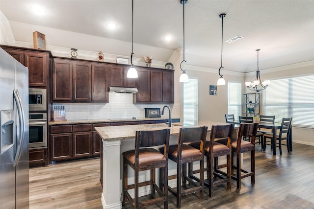 kitchen with appliances with stainless steel finishes, a breakfast bar, decorative light fixtures, an island with sink, and light stone counters