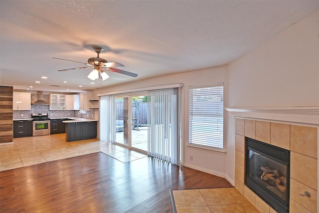 kitchen with a tiled fireplace, hardwood / wood-style flooring, ceiling fan, wall chimney range hood, and stainless steel range oven