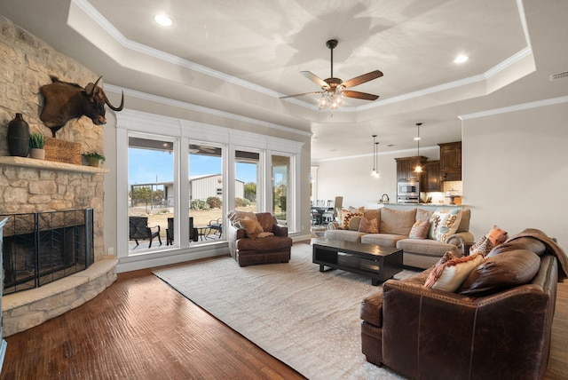 living room featuring ornamental molding, a raised ceiling, ceiling fan, wood-type flooring, and a stone fireplace