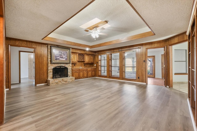 unfurnished living room featuring a stone fireplace, light hardwood / wood-style flooring, ceiling fan, a textured ceiling, and a tray ceiling