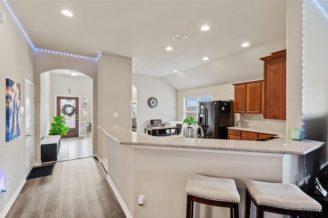 kitchen featuring a kitchen bar, black fridge with ice dispenser, kitchen peninsula, and light stone counters