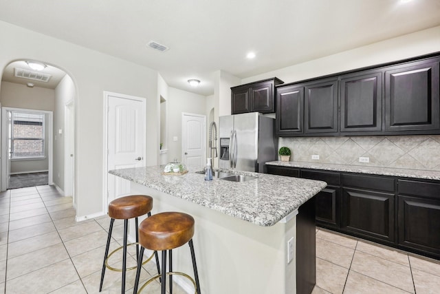 kitchen with a kitchen island with sink, stainless steel refrigerator with ice dispenser, tasteful backsplash, light tile patterned flooring, and light stone counters