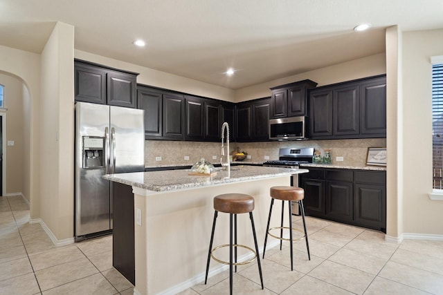 kitchen with a kitchen island with sink, sink, light stone countertops, appliances with stainless steel finishes, and light tile patterned flooring