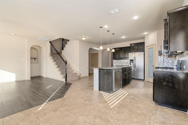 kitchen featuring a center island with sink, backsplash, stainless steel refrigerator with ice dispenser, and hanging light fixtures