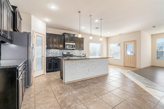 kitchen with a kitchen island with sink, tasteful backsplash, light tile patterned flooring, and appliances with stainless steel finishes