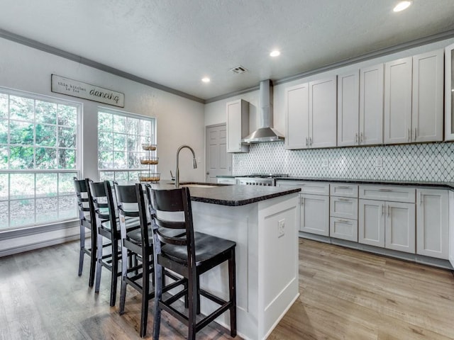 kitchen with a kitchen breakfast bar, sink, wall chimney exhaust hood, light wood-type flooring, and an island with sink