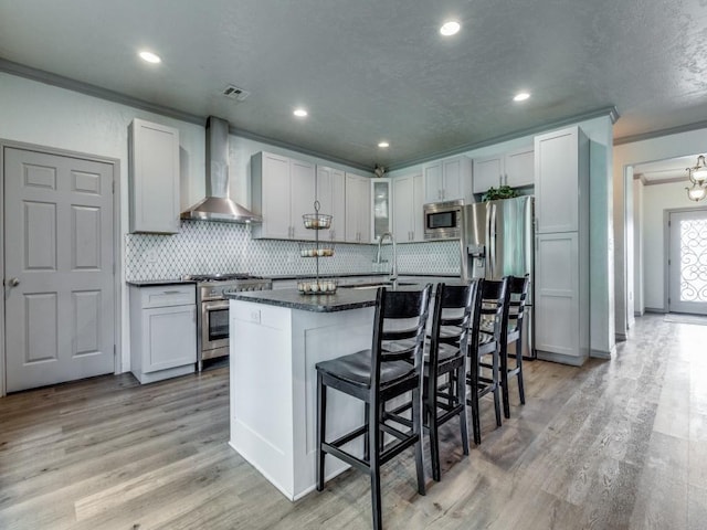 kitchen with a kitchen breakfast bar, ornamental molding, wall chimney exhaust hood, stainless steel appliances, and a kitchen island with sink