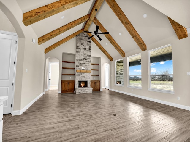 unfurnished living room featuring beam ceiling, built in shelves, ceiling fan, a stone fireplace, and high vaulted ceiling