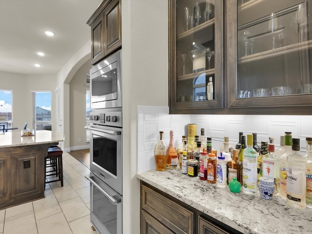 bar with light stone countertops, appliances with stainless steel finishes, dark brown cabinets, and light tile patterned floors