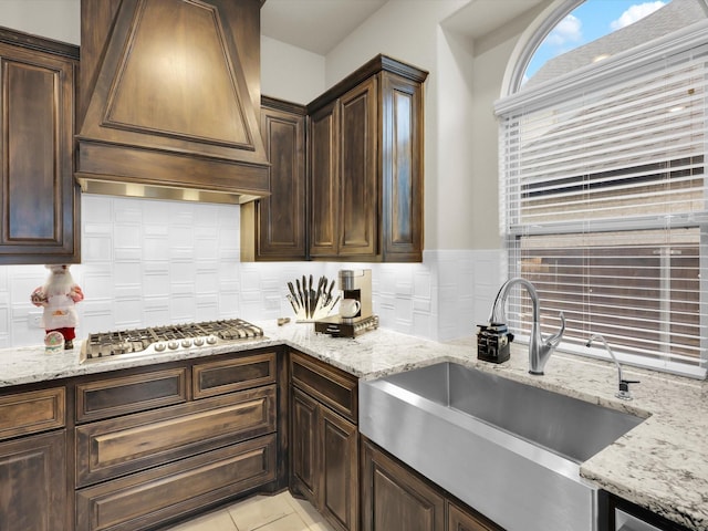 kitchen with custom range hood, a sink, dark brown cabinetry, light stone countertops, and stainless steel gas cooktop