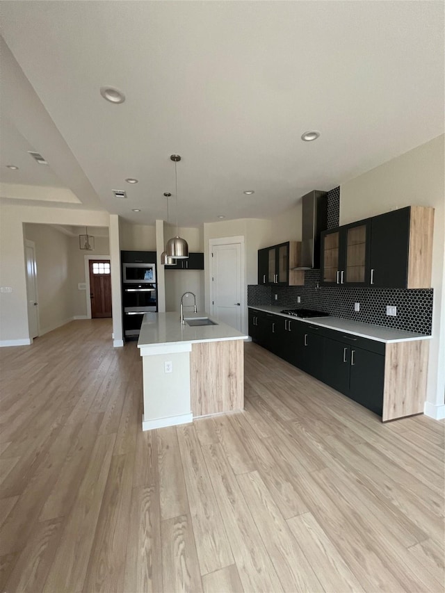 kitchen featuring sink, wall chimney range hood, pendant lighting, a kitchen island with sink, and light wood-type flooring