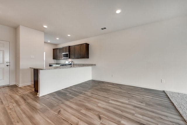 kitchen with kitchen peninsula, appliances with stainless steel finishes, light stone counters, dark brown cabinets, and light hardwood / wood-style flooring