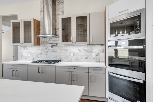 kitchen with wall chimney exhaust hood, gray cabinets, stainless steel appliances, light countertops, and backsplash