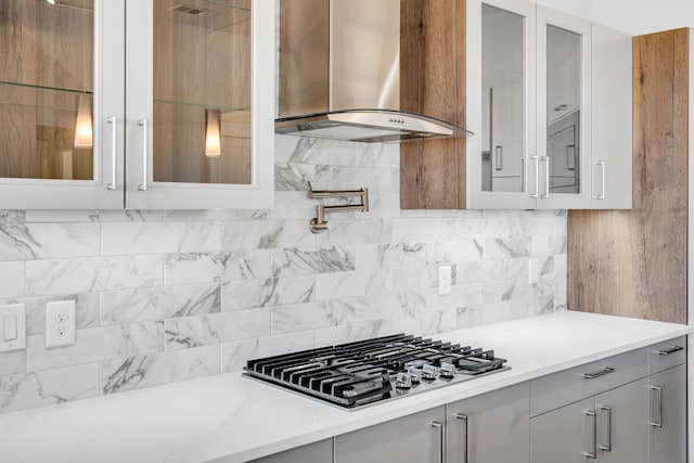 kitchen featuring wall chimney exhaust hood, light stone countertops, gray cabinetry, stainless steel gas stovetop, and backsplash